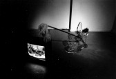 A black and white photograph of a mop on top of a working CRT television in a gallery setting.