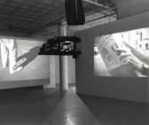 A black and white photograph with two projectors hanging central with projections on one wall and another hanging screen in a gallery space.