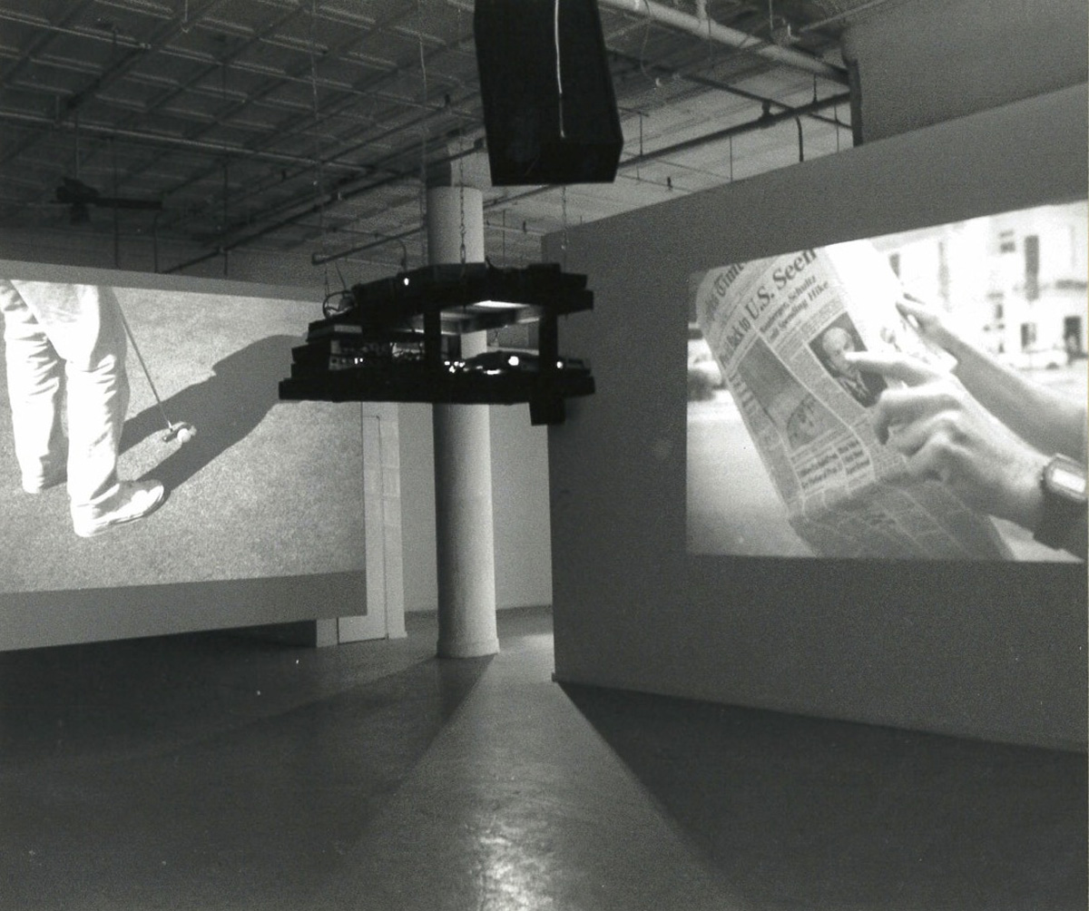 A black and white photograph with two projectors hanging central with projections on one wall and another hanging screen in a gallery space.