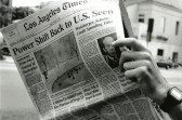 A black and white image of a hand holding a newspaper with a view of the street and surroudning buildings in the background.