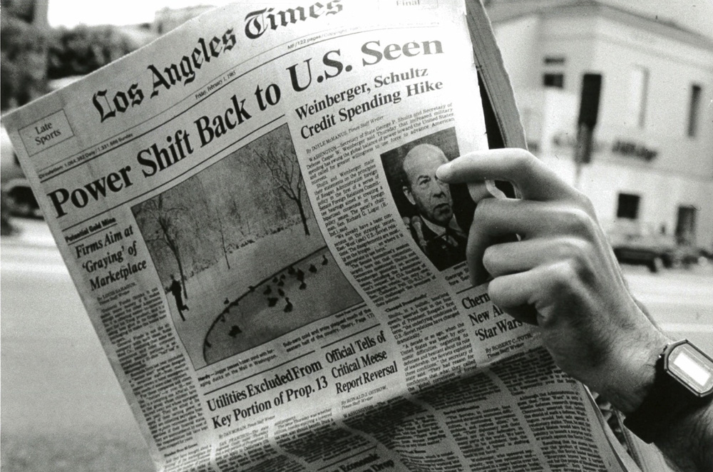 A black and white image of a hand holding a newspaper with a view of the street and surroudning buildings in the background.