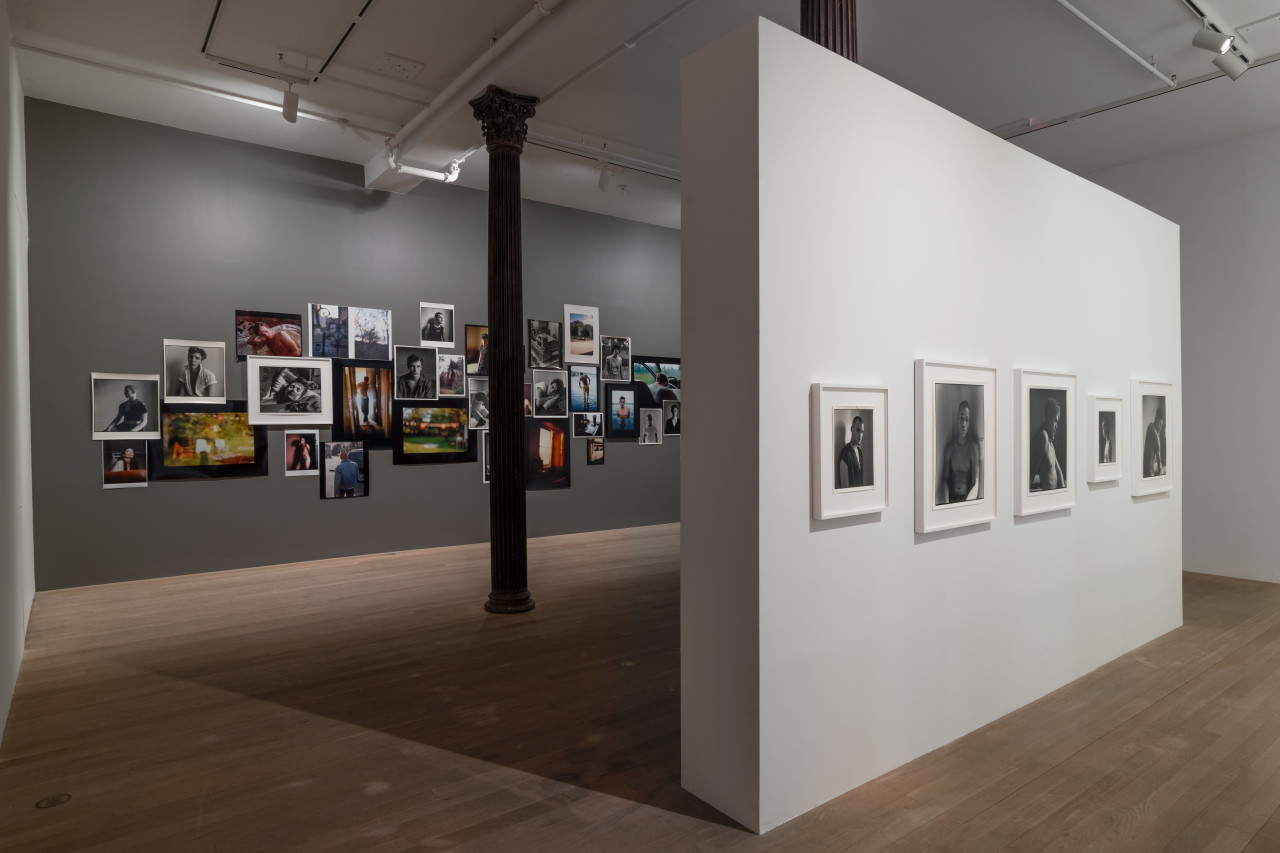 On the back wall, painted in grey, a collage of photographs intertwines and overlaps. Directly in front, a white gallery wall displays five black and white portraits of various sizes.