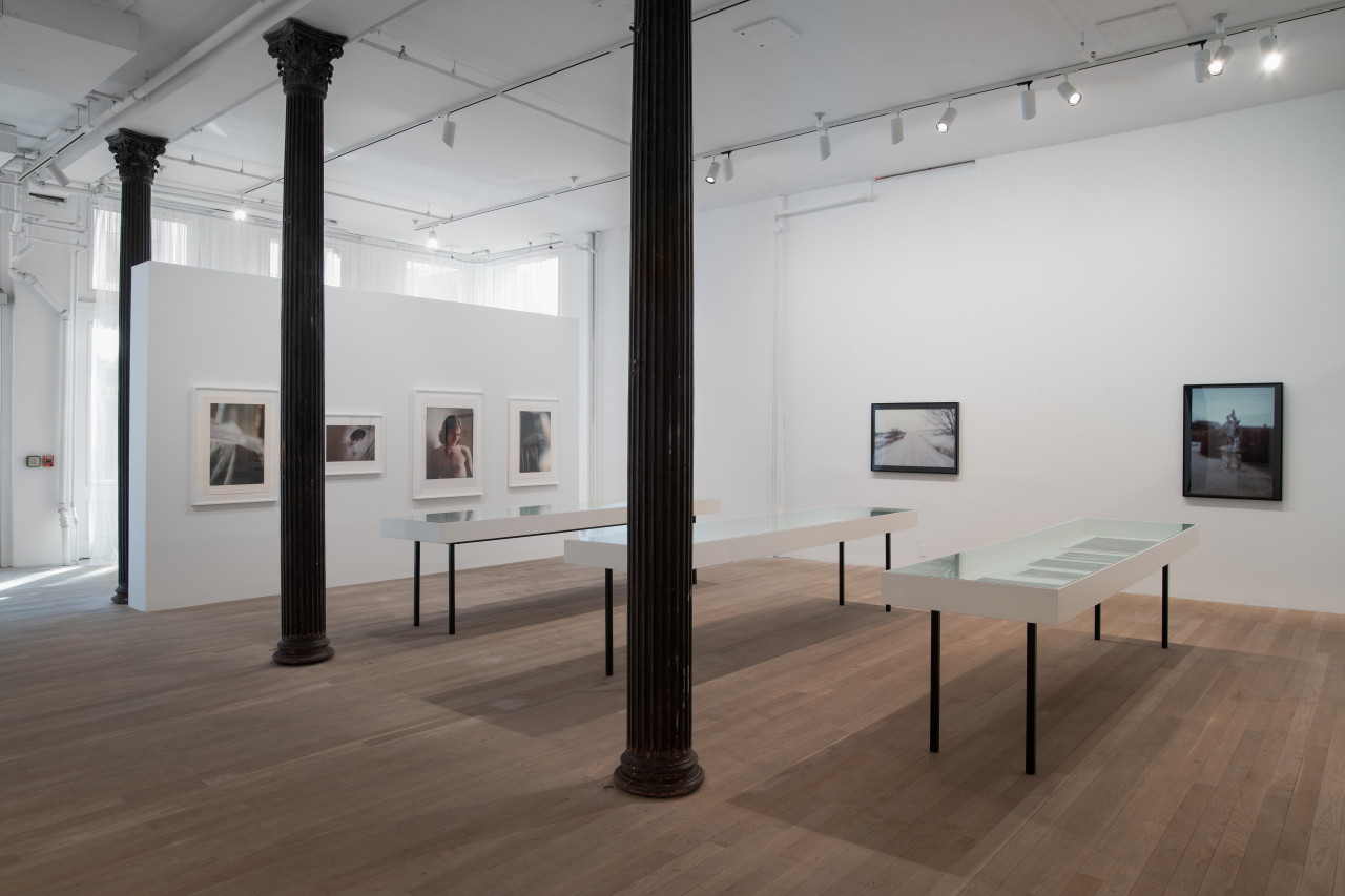 In an open gallery space, three vitirnes stand surrounded by walls displaying large color photographs.