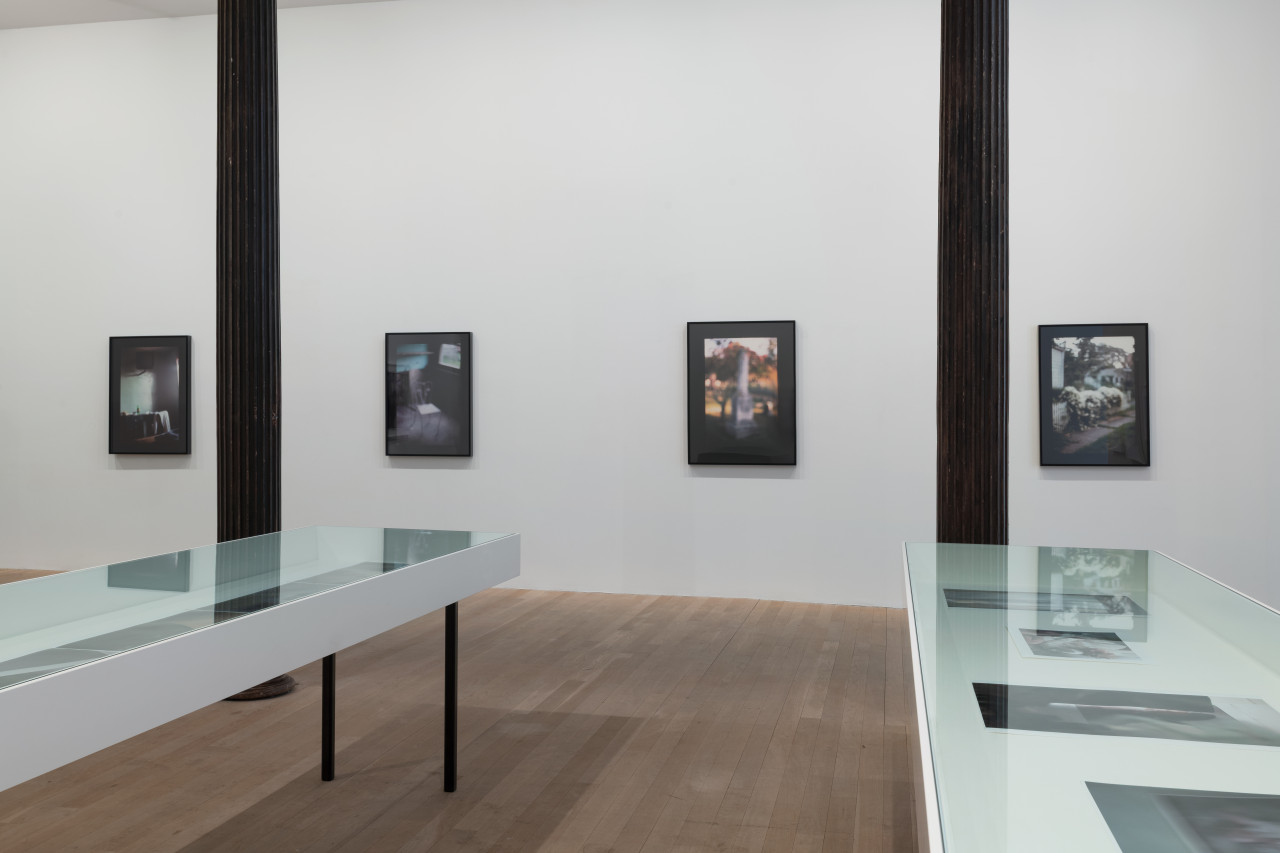 Two glass-topped vitirines reveal photographs of various sizes. Behind, four large color photographs with thick black borders hang on the gallery wall.