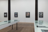 Two glass-topped vitirines reveal photographs of various sizes. Behind, four large color photographs with thick black borders hang on the gallery wall. 