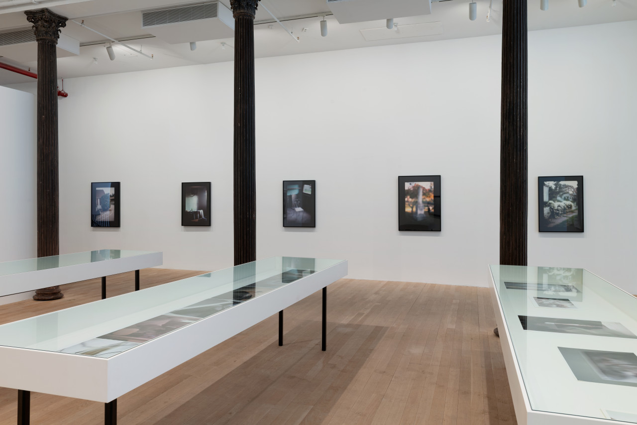 Three glass topped vintrines carry smaller color photographs. The gallery walls behind display five photographs, each depicting a different hazy space adorned with thick black borders.