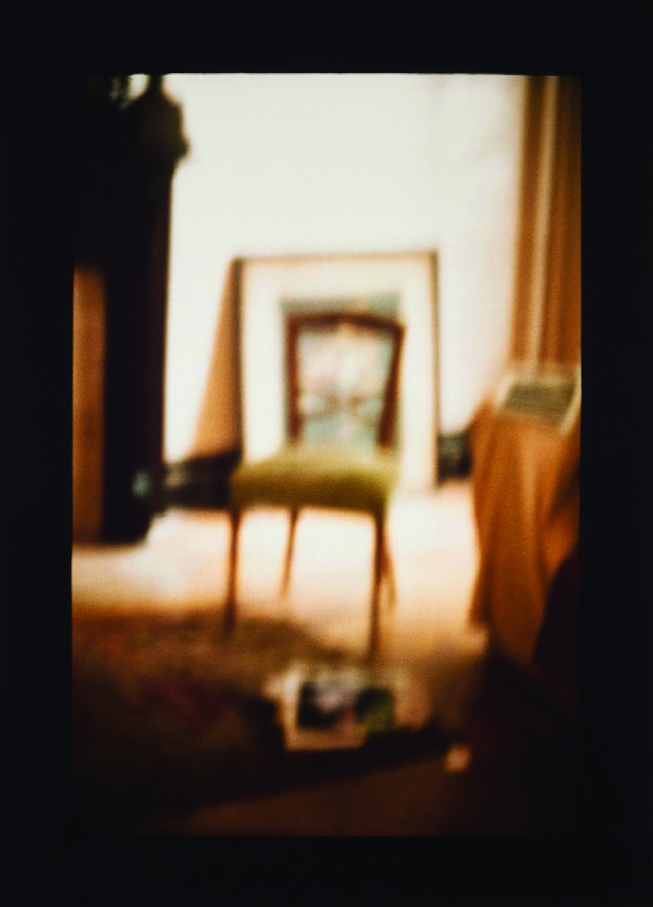 An out-of-focus photograph of a wooden chair in a golden-lit room decorated with curtains, a carpet, and a framed artwork leaning against the back wall.