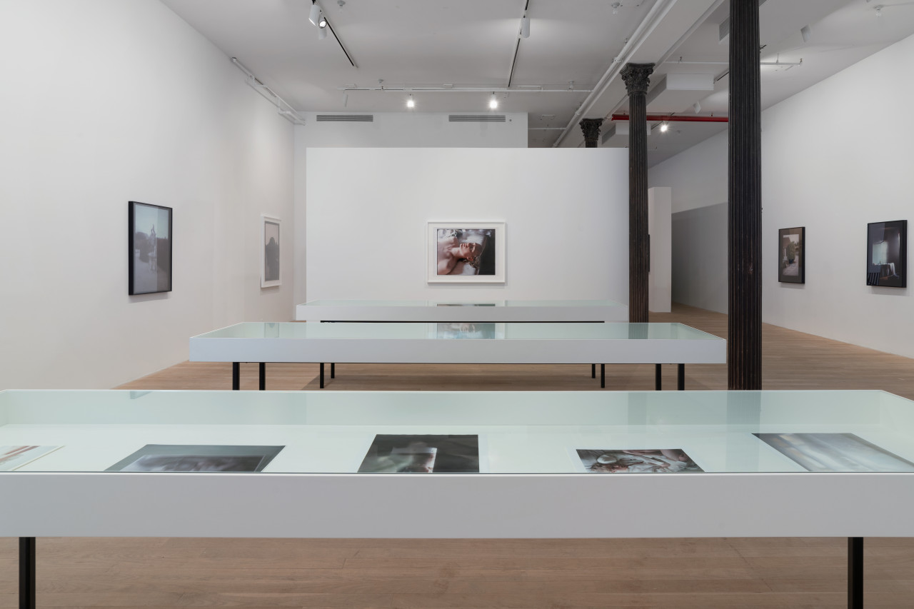 In an open gallery space, three vitirnes stand surrounded by walls displaying large color photographs with thick black borders. The inner display of a vitrine reveals smaller color photographs.