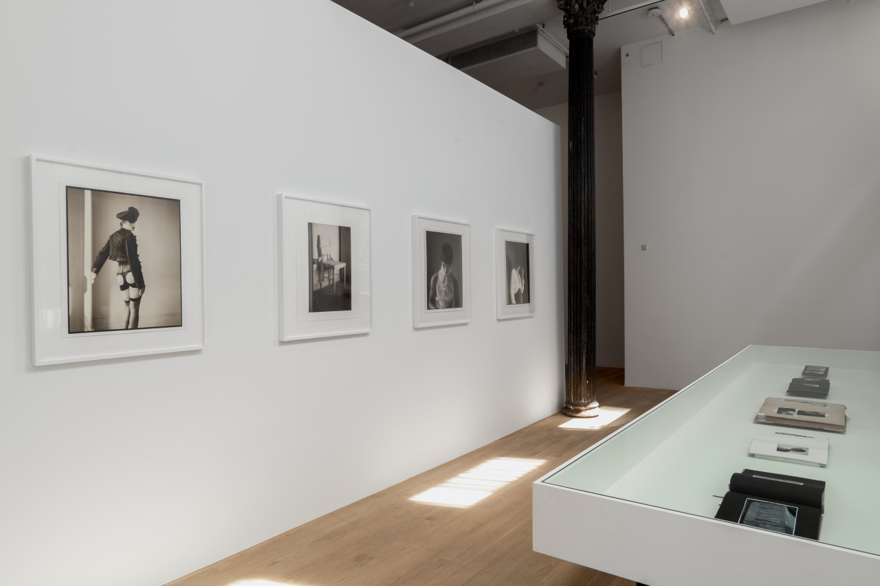 A vitrine and a gallery wall are parallel in the bright gallery space. The vitrine displays books of different sizes opened to various pages. On the wall, four black and white photographs hang vertically in sequence.