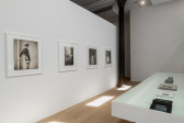 A vitrine and a gallery wall are parallel in the bright gallery space. The vitrine displays books of different sizes opened to various pages. On the wall, four black and white photographs hang vertically in sequence. 