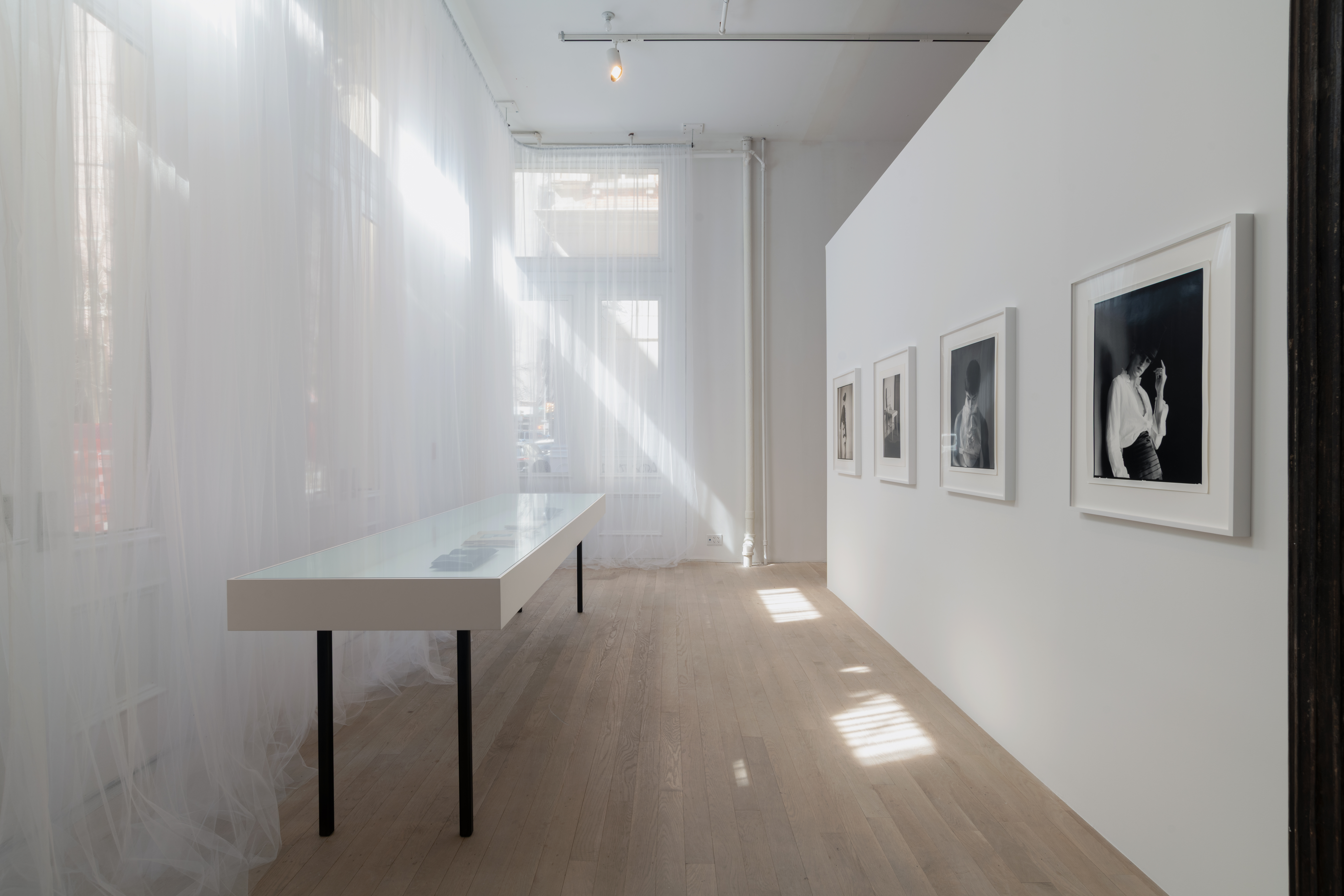 Toile curtains gather around the gallery window, producing a soft, glowing white light. Four black and white photographs hang across from the windows, receding into space. 