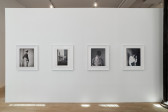 Four black and white photographs hang on a gallery wall. The floor is speckled with light, and the photographs, mainly portraits, are dramatic yet soft.  