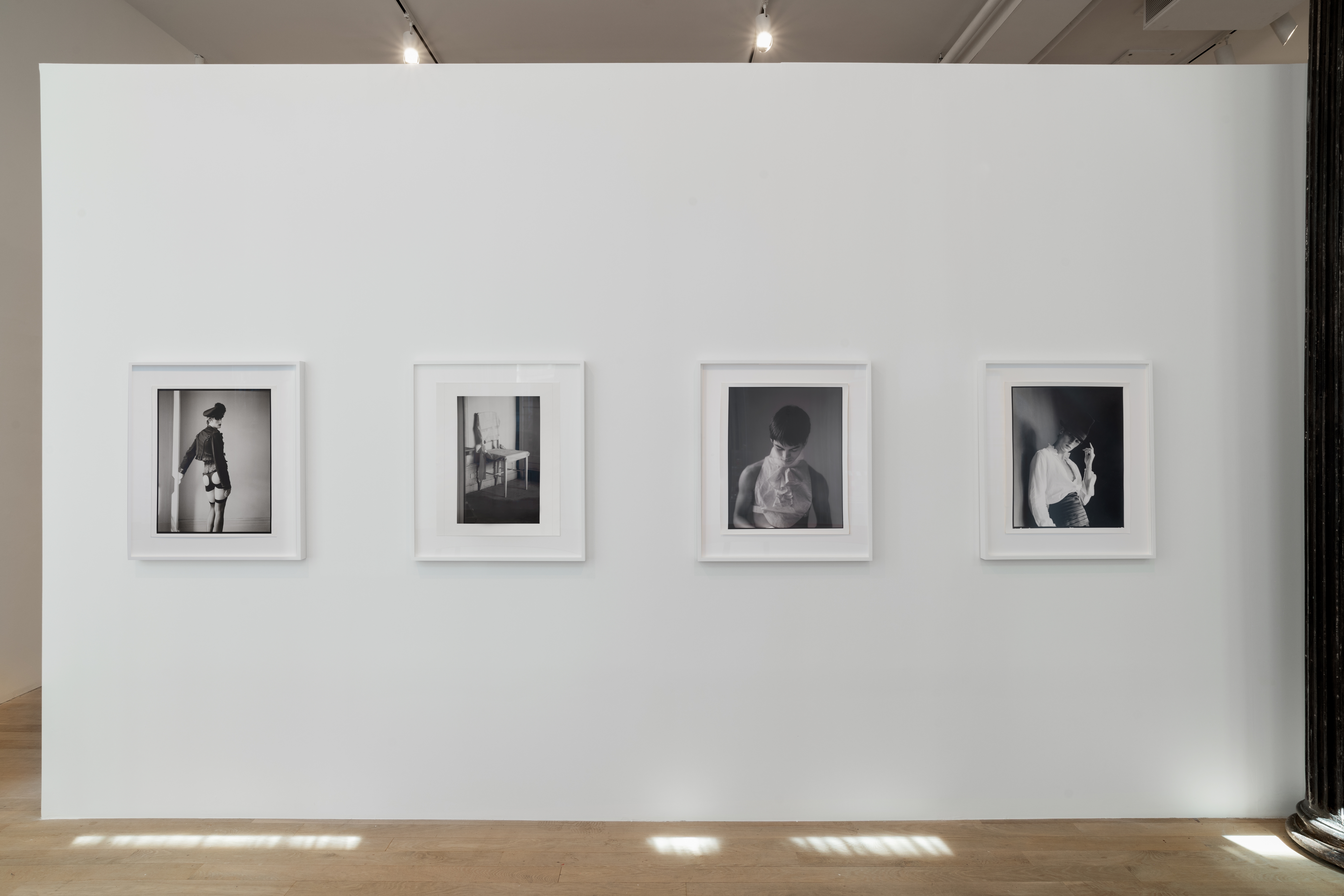 Four black and white photographs hang on a gallery wall. The floor is speckled with light, and the photographs, mainly portraits, are dramatic yet soft.  