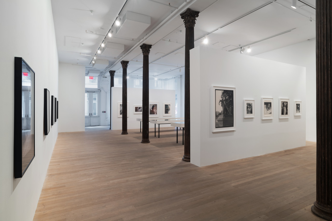 An open gallery space shows various photographs hung in sequence. The wall closest in proximity displays black and white images. Behind sit vitrines, followed by another wall of color images.