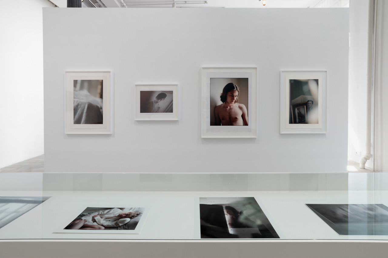 The inner display of a vitrine reveals four smaller color photographs. Directly above on a bright gallery wall, hang four larger photographs. One of the four is a slightly larger-than-life portrait of a young man printed in soft tones.