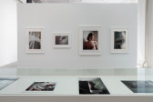 The inner display of a vitrine reveals four smaller color photographs. Directly above on a bright gallery wall, hang four larger photographs. One of the four is a slightly larger-than-life portrait of a young man printed in soft tones. 