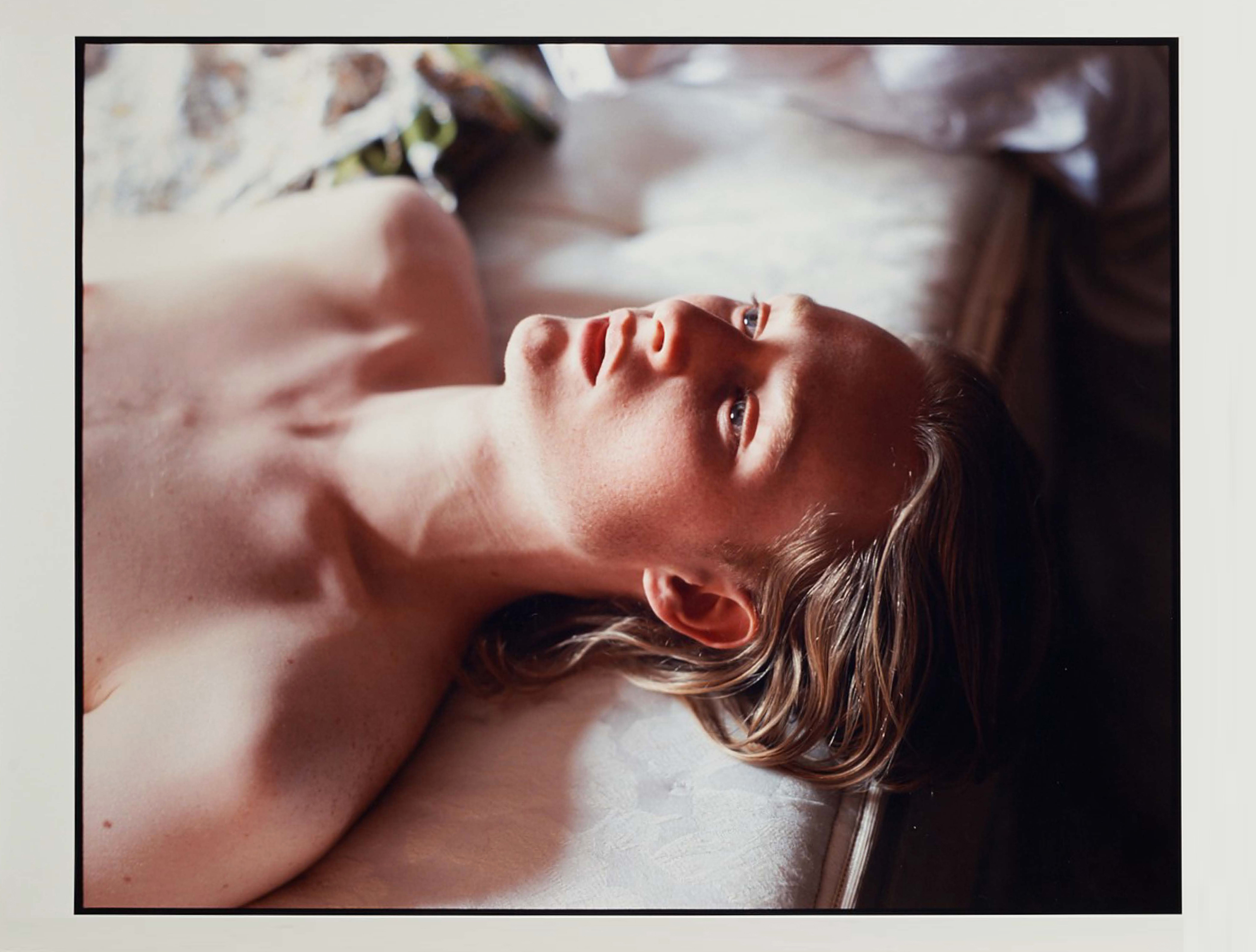 A figure lays face up on a white mattress. His expression is languid and his hair falls over the edge of the mattress.