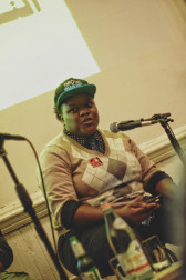 Marz Saffore, dressed in a beige argyle swearer, a polka dot button up, jeans, and a green cap, sits and speaks into a microphone.