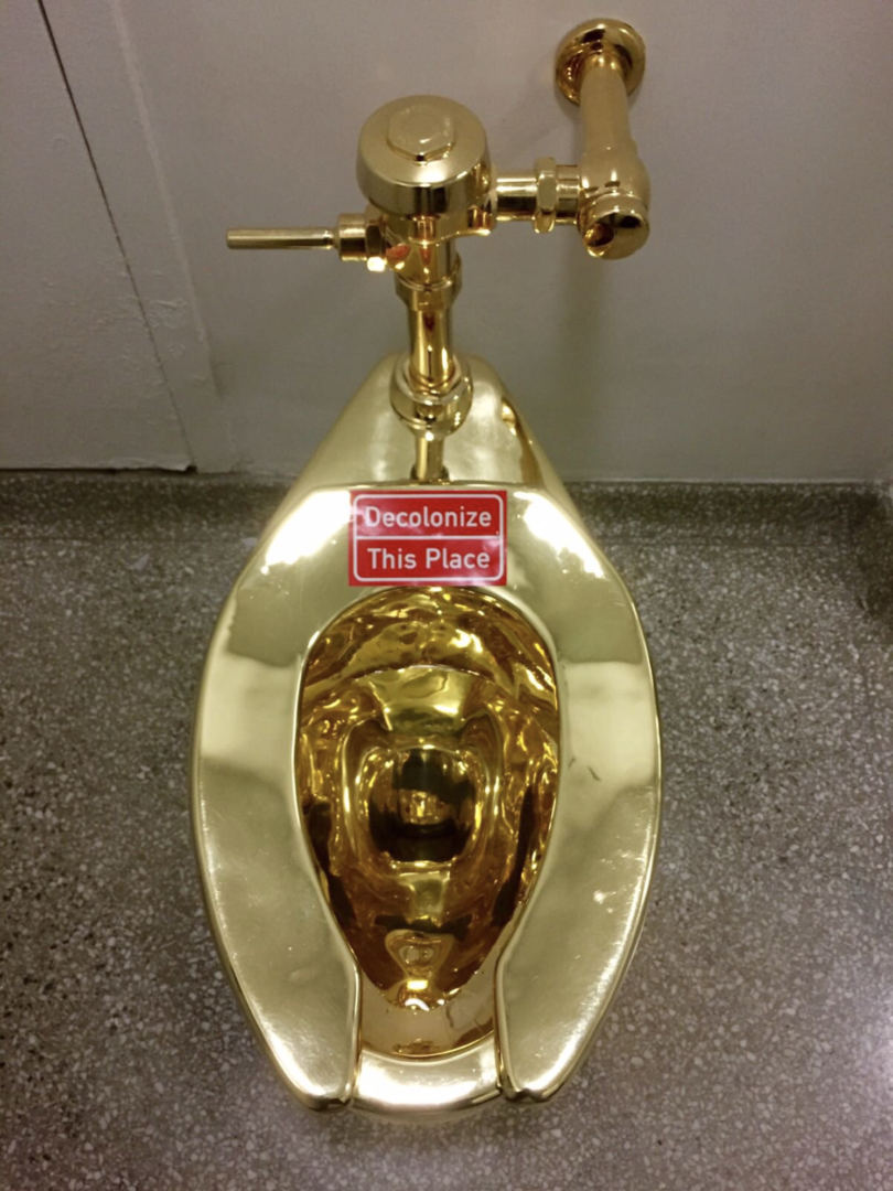 An image of a gold public bathroom toilet with a red Decolonize This Place sticker placed at the top of the toilet seat.
