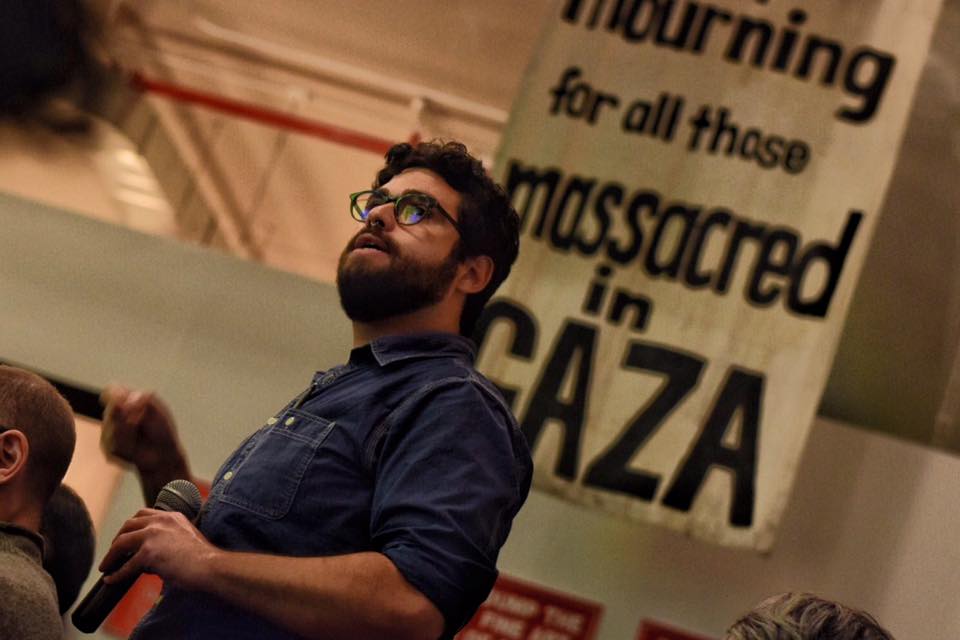 An upwards-angeled view of an audience member, wearing a dark denim shirt, with a poster in the background that reads 