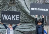 Two children holding up black signs with white text in front of a building covered by an opaque plastic material, one sign reads 