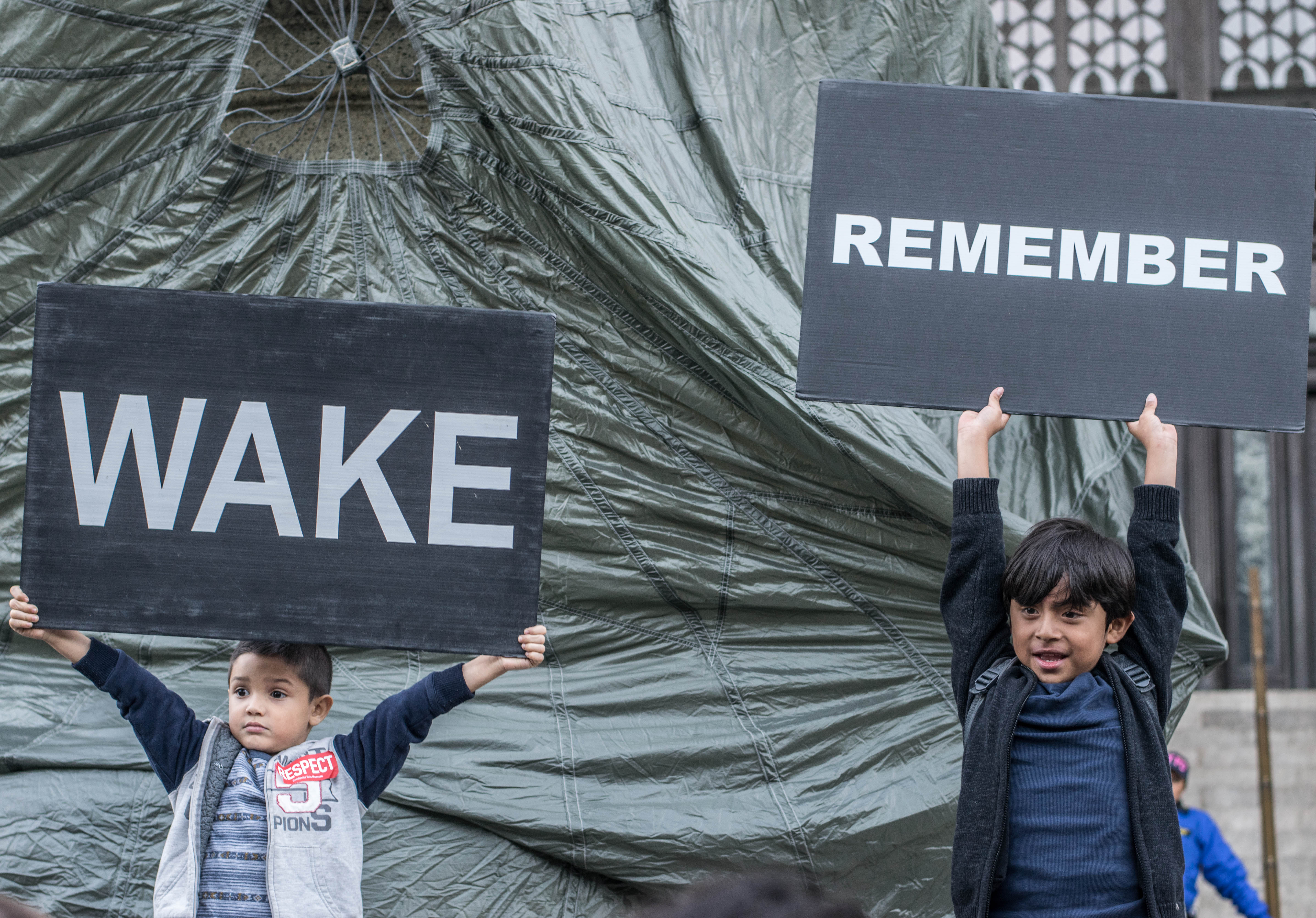 Two children holding up black signs with white text in front of a building covered by an opaque plastic material, one sign reads 