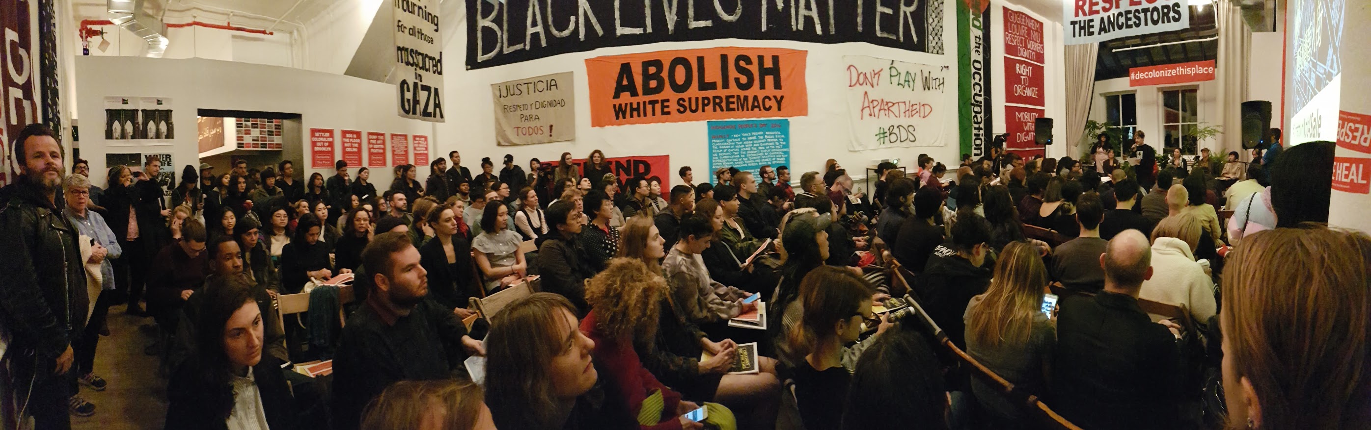 A panoramic image of the audience at Artists Space and Decolonize This Place's event, featuring colorful banners on the wall that read 