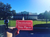 A person's hand holding up a Decolonize This Place sticker in front of the White House, with a security guard visible in the background.