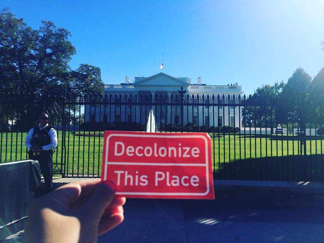 A person's hand holding up a Decolonize This Place sticker in front of the White House, with a security guard visible in the background.