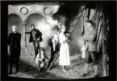 Seven people are in a courtyard in period dress, facing the camera.  Some stand on a ladder, another leans against a colonnade in the background.  Smoke billows out behind them from an unseen source.
