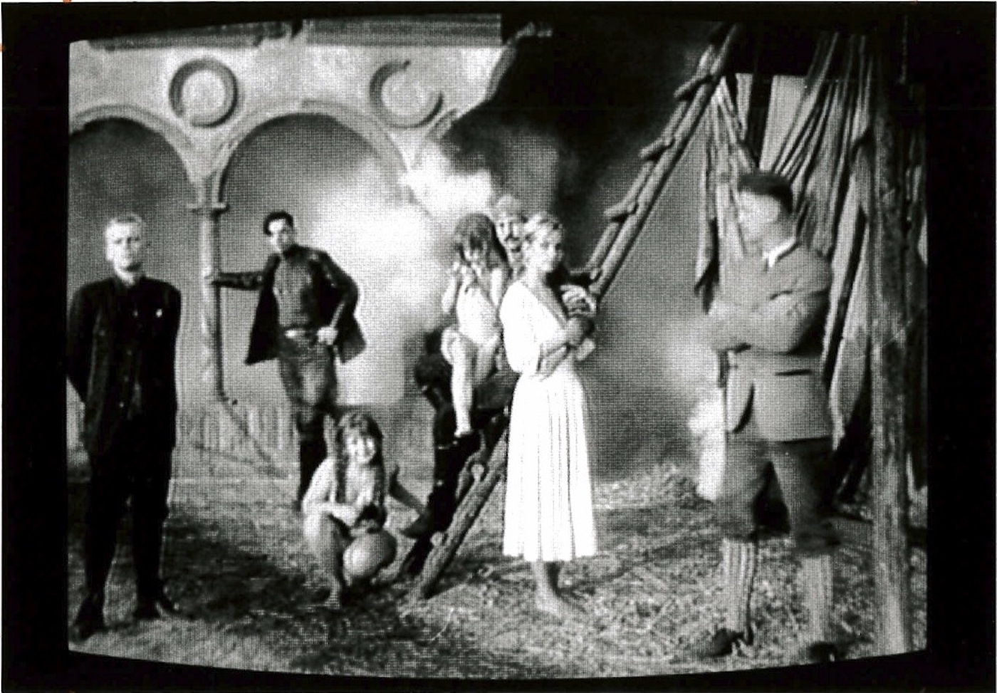 Seven people are in a courtyard in period dress, facing the camera.  Some stand on a ladder, another leans against a colonnade in the background.  Smoke billows out behind them from an unseen source.