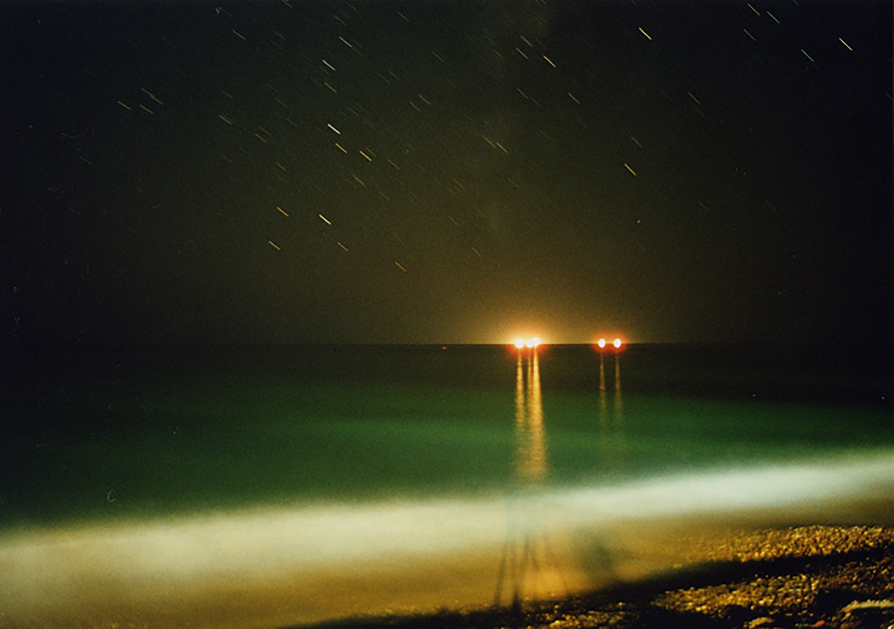 An outdoor landscape at night. Raindrops fall from the sky, and several dots of bright light are visible on the horizon over a great expanse of water, reflecting yellow light onto the water's surface.