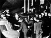A dense crowd of people surrond a casket being carried into the back of a hearse.
