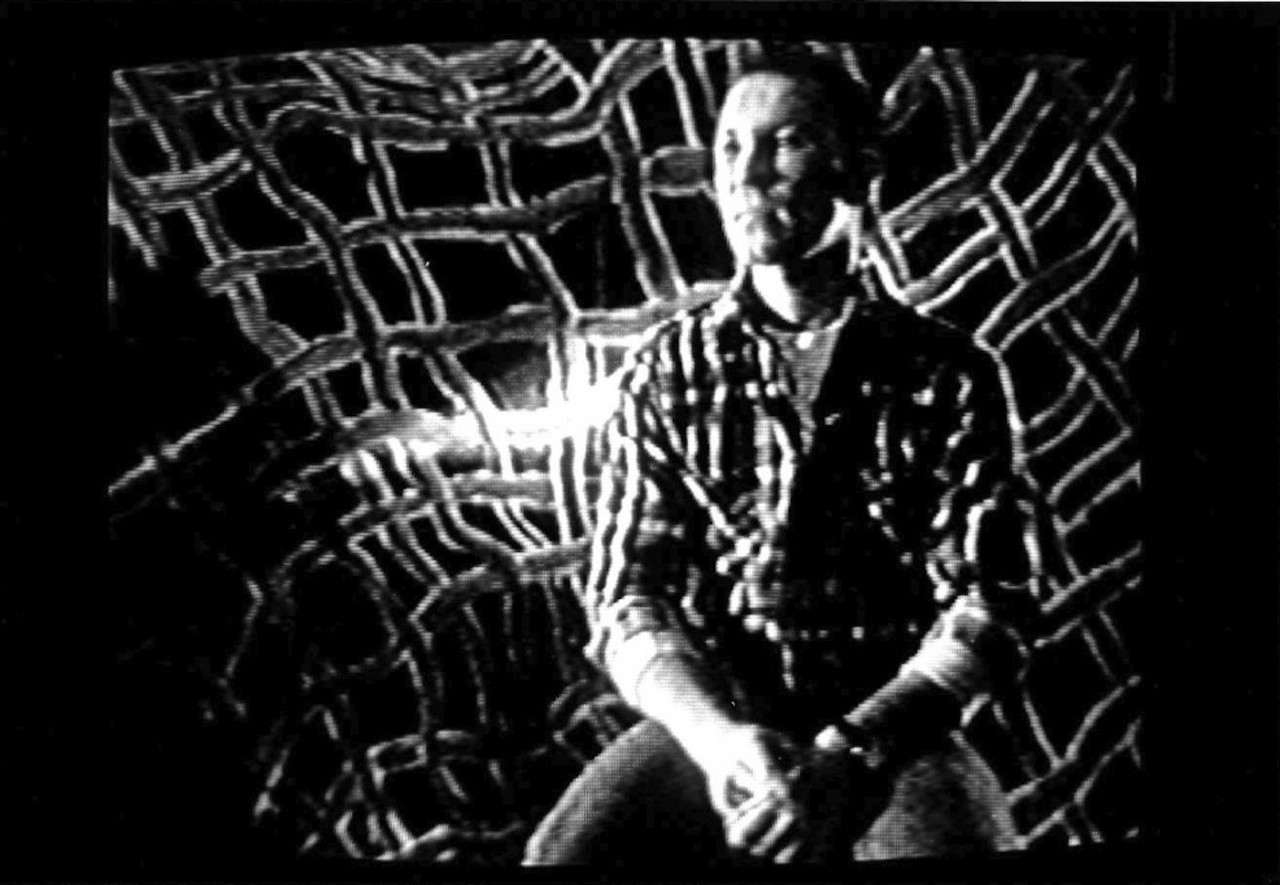 A person in a plaid shirt and jeans is seated with their hands in their lap in from of a plaid background, looking to the left of the camera.