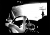 A person driving a car is seen from behind, as if the camera is in the backseat.  Their face is hidden by the headrest of the car seat.  Trinkets hang from the rearview mirror.