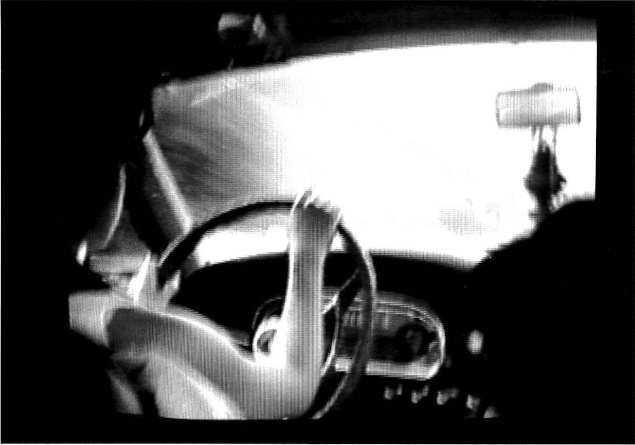 A person driving a car is seen from behind, as if the camera is in the backseat.  Their face is hidden by the headrest of the car seat.  Trinkets hang from the rearview mirror.