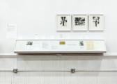 A bookcase displays documents within as well as on top of its glass. Above, right of center, three black and white photographs are hung, in white frames, on the wall.