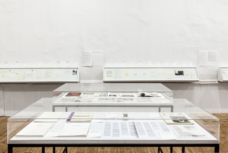 A view of two vitrines, one in front of the other, displaying documents in front of a book case on the wall. Documents containing text are hung on the wall.