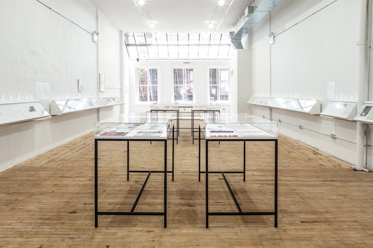 A view of five vitrines, displaying various documents, arranged in columns in the middle of a room. On either wall, book cases used for display.