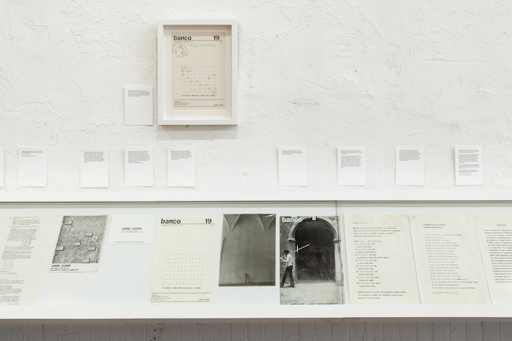 A book shelf displays paper documents containing black and white images. Above, smaller documents, and a similar paper displayed in a white frame.
