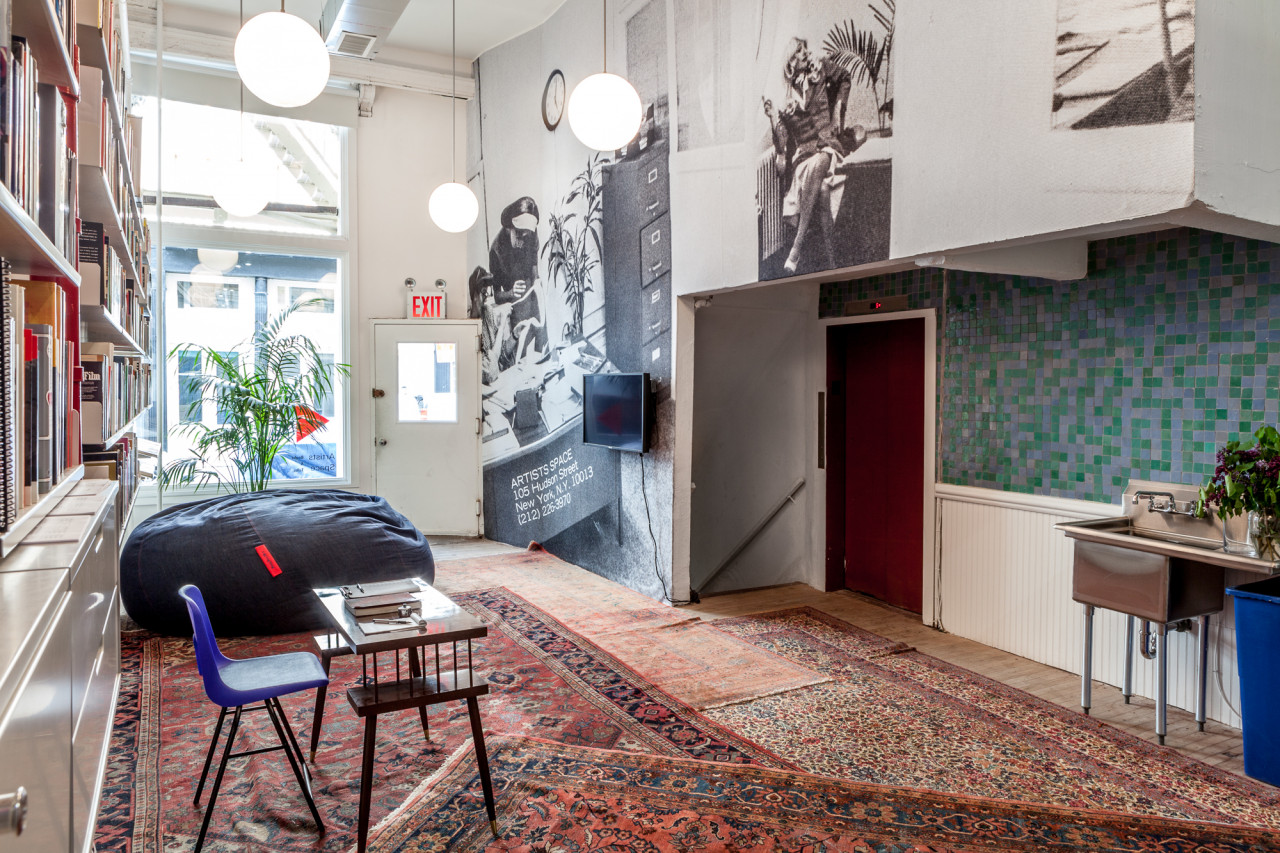 An entryway with layered, decorative carpets and black-and-white images displayed across the height and length of the wall. A small school desk sits on the left in front of a large bookshelf.