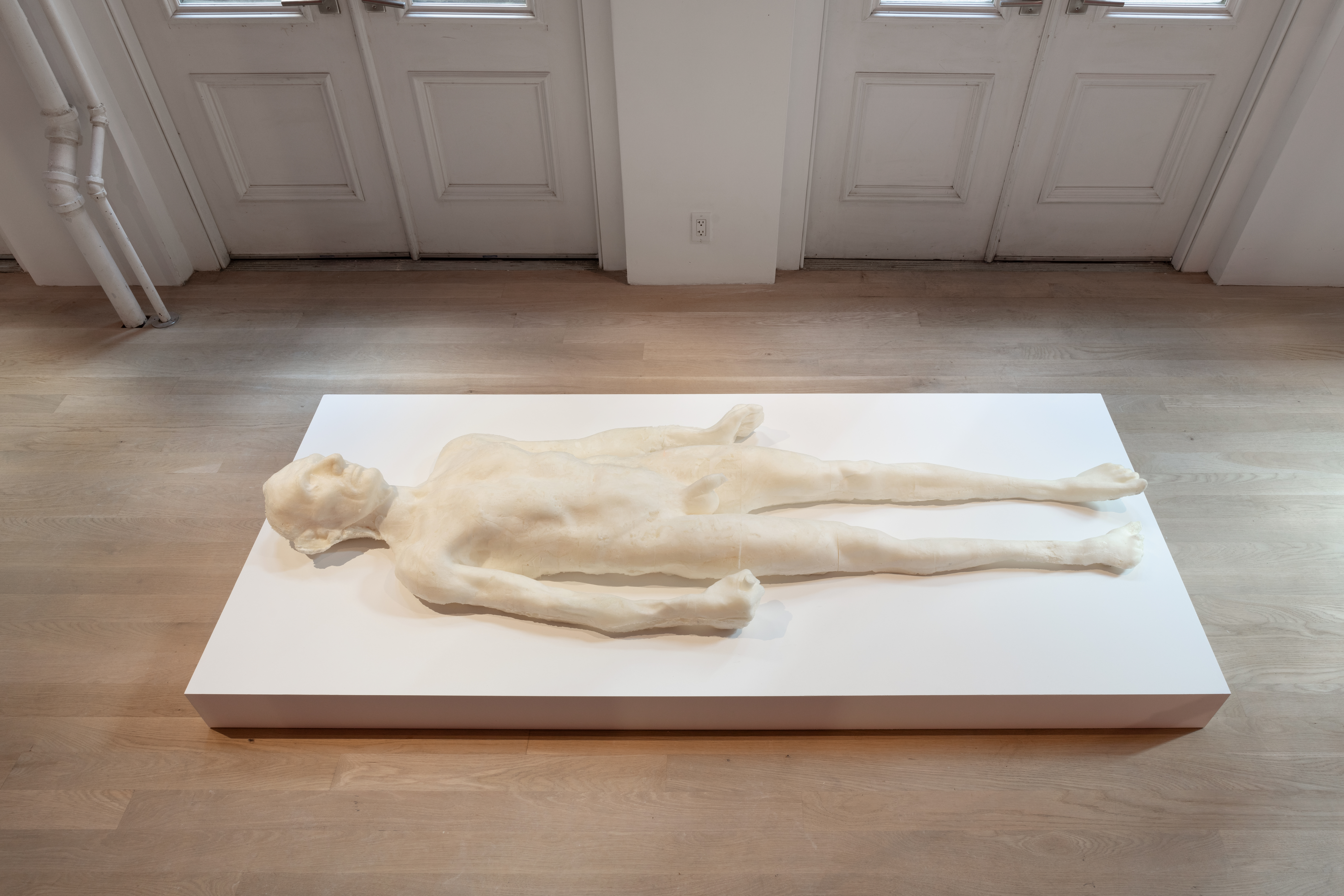 A life-size off-white foam sculpture of a naked human form, in the likeness of Abraham Lincoln, rests supine on a low white platform.