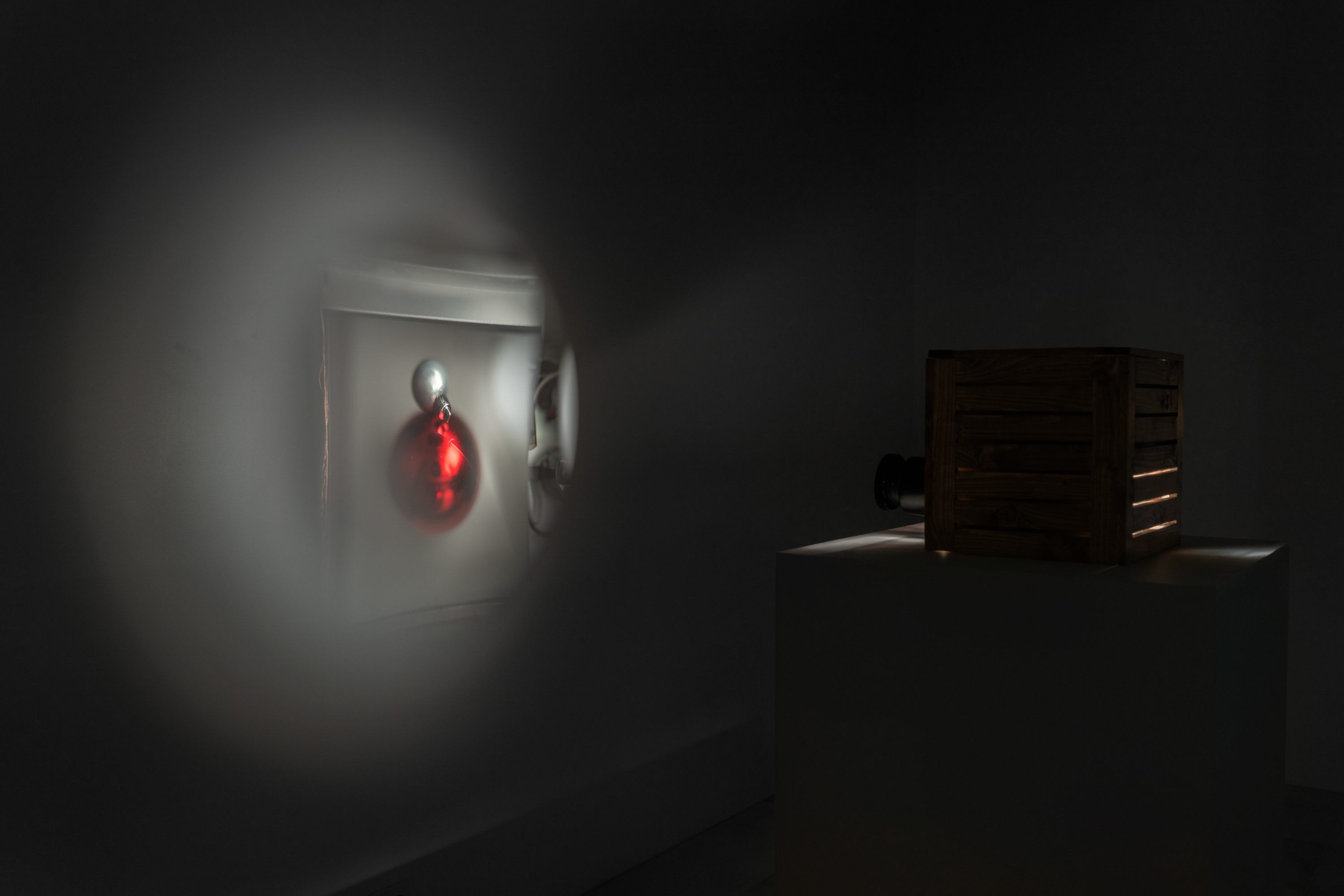 Color image of a small pinhole projector, sitting on a pedestal and facing a wall to the left of the image. On the wall, an image of a red pearlescent ball, with a small white pearlescant ball attached on top, is projected.