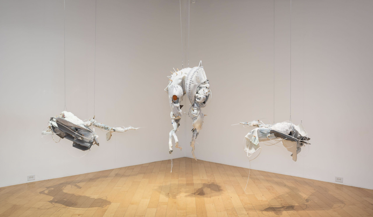 Color image of the corner of a gallery space with three hanging sculptures. The sculptures are made of pigmented silicone, old clothes, zip ties and various other found materials. The sculptures are mainly made of white materials, and feature anthropormorphic and abstracted forms.