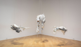 Color image of the corner of a gallery space with three hanging sculptures. The sculptures are made of pigmented silicone, old clothes, zip ties and various other found materials. The sculptures are mainly made of white materials, and feature anthropormorphic and abstracted forms.