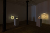 Color image of the corner of a darkened gallery space with two small pinhole projector sculpture installations. The pinhole projector on the left is housed in a polka-dot patterned gift-wrapped box. On the wall, an image of a white ceramic teacup with a radial swirl pattern is projected. On the right, the projector is wrapped in white foam padding and secured with red string. On the wall, it projects an image of a square containing an golden metal hoop and pin along with four small, glittering pink and green strawberry ornaments.