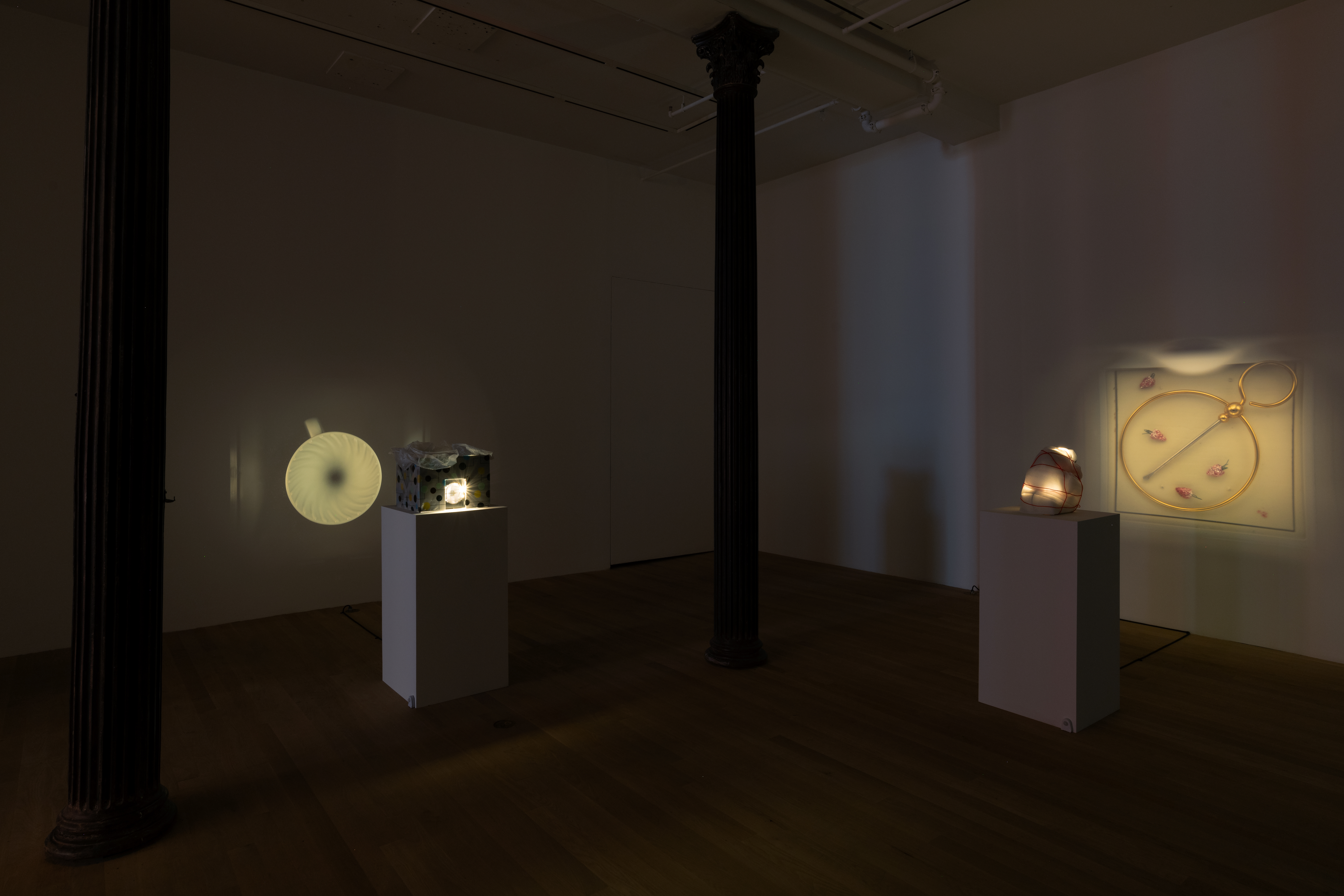 Color image of the corner of a darkened gallery space with two small pinhole projector sculpture installations. The pinhole projector on the left is housed in a polka-dot patterned gift-wrapped box. On the wall, an image of a white ceramic teacup with a radial swirl pattern is projected. On the right, the projector is wrapped in white foam padding and secured with red string. On the wall, it projects an image of a square containing an golden metal hoop and pin along with four small, glittering pink and green strawberry ornaments.