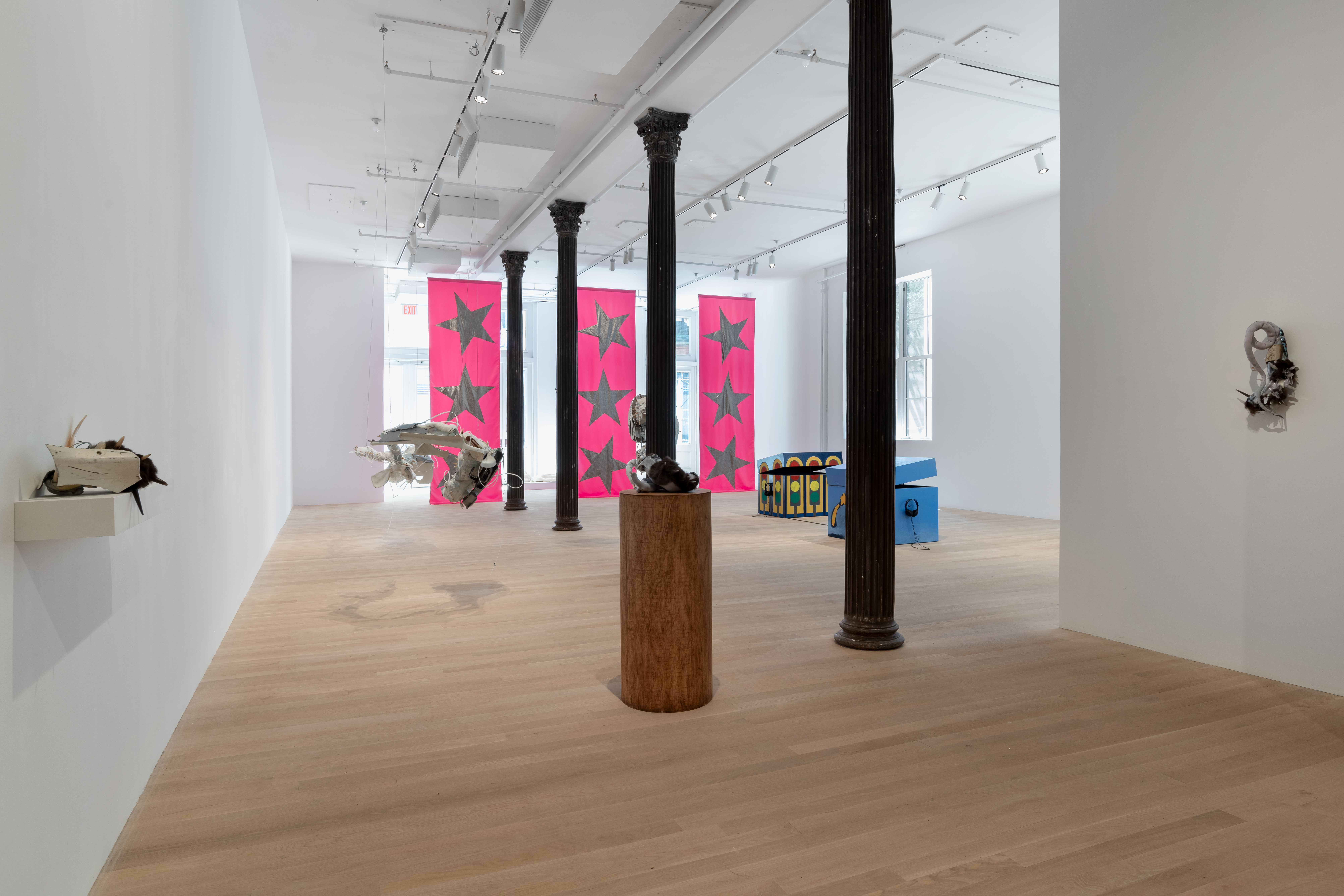 A receding row of columns lines our view into the far gallery space. In the foreground, several of Ruben Ulises Rodriguez Montoya's biomorphic sculptures are displayed on walls, resting on a pedestal, or hanging from the ceiling. In the distance, two of Matthew Langan-Peck's monumental painted toy box sculptures sit on the gallery floor; a set of headphones hangs on the side of each box. In the background, Isabelle Frances McGuire's three large pink banners emblazoned with silver stars hang from ceiling to floor.
