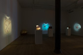 Color image of a darkened gallery installation with multiple projectors and a golden egg sculpture in the left back corner. In the left foreground, a pinhole projector wrapped in foam and secured with red string sits on a pedestal. On the wall to the left, an image of a golden ornamental hoop and pin, along with four small pink and green strawberries, is projected. In the center back, a pinhole project inside a rectangular plaid plastic travel bag with straps sits atop a pedestal. It projects an image of a green square with a crenellated metal opening onto the back wall. To the right, a projector inside a plywood box sits atop a pedestal. On the wall to the right, an image of an image of a red pearlescent ball, with a small white pearlescant ball attached on top, is projected.
