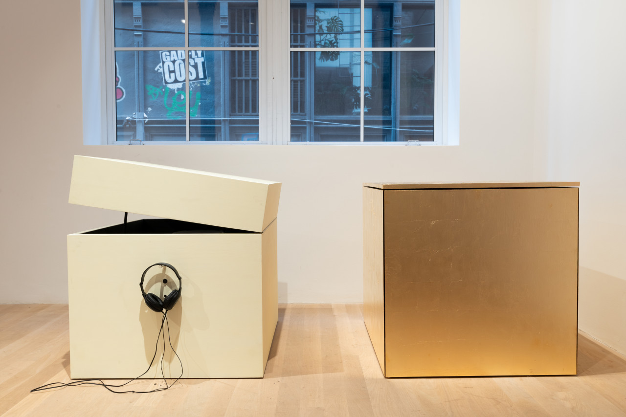 Closeup view of two wooden box sculptures sitting side by side in a gallery. The box on the left is painted light yellow and has a pair of headphones hanging from its side; its lid is slightly ajar. The closed box on the right is covered completely in imitation gold leaf.